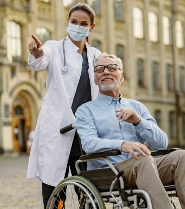 Full length shot of caring young nurse in protective mask taking care of senior man handicapped